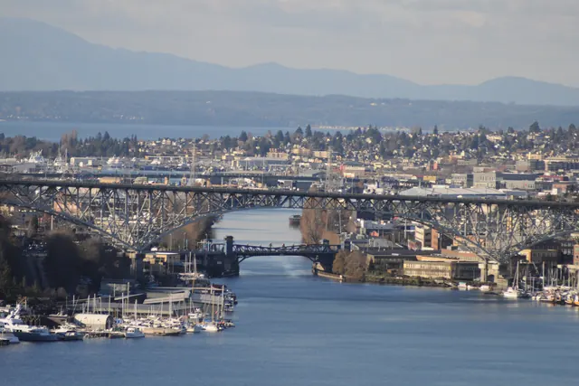 The Fremont Cut lined with moored boats, passing under the tall arch of the Aurora Bridge and the low Fremont Bridge, with the Olympic Mountains in the distance.