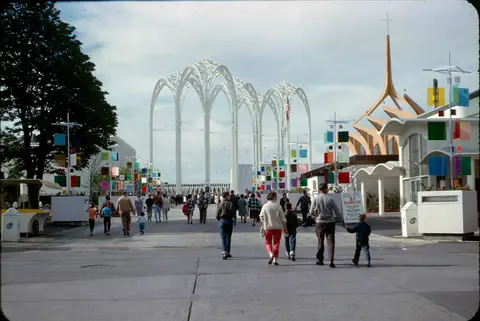 Families walking across a wide plaza toward tall white lattice arches, with colourful banners and kiosks.
