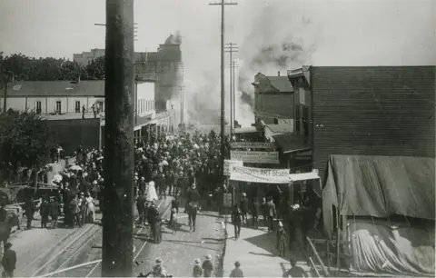 A crowded dirt street of wooden buildings and tents, with thick smoke rising from burning buildings in the distance.