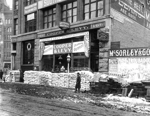 A brick storefront signed Cooper and Levy Pioneer Outfitters, with a long wall of stacked flour sacks and crates on the sidewalk.