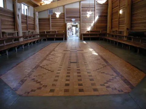 A long hall with warm cedar plank walls and posts, benches along both sides and a patterned wooden floor.