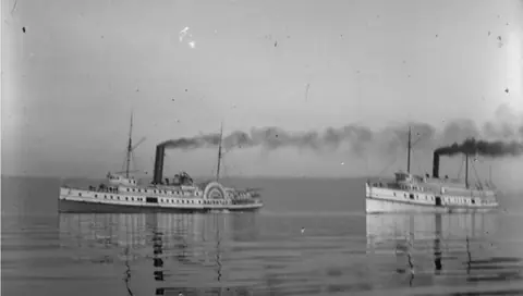 Two white steamboats with tall black smokestacks racing side by side across glassy water.