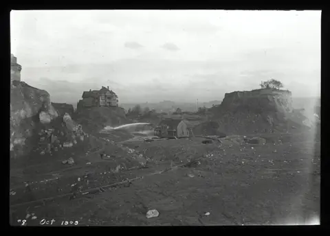 A cut-away hillside of bare earth with two houses stranded on tall mounds, and a jet of water arcing across.