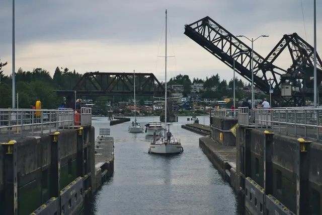 A white sailboat in a narrow concrete lock chamber, with a raised black steel railway bridge behind it and green hills beyond.