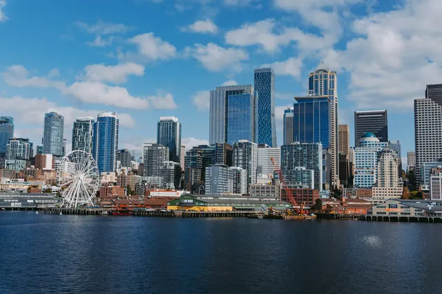 Downtown towers under a blue sky across dark blue water, with the white Ferris wheel and the low piers along the shore.